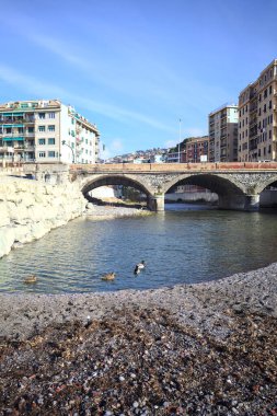 Güneşli bir günde, nehrin çıkışında kurumuş bir yerden görülebilen, şehrin binalarıyla çevrili bir nehrin üzerindeki kemerli alçak köprü.