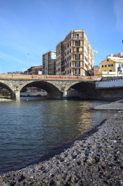 Güneşli bir günde, nehrin çıkışında kurumuş bir yerden görülebilen, şehrin binalarıyla çevrili bir nehrin üzerindeki kemerli alçak köprü.