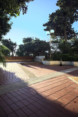 Tiled balcony partially in the shade in a promenade bordered by flowerbeds filled with trees on a sunny day