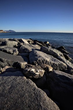 Tip of a promontory with buildings and a cliff away in the distance seen from a small cove and framed by rocks of a breakwater