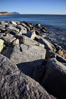 Tip of a promontory with buildings and a cliff away in the distance seen from a small cove and framed by rocks of a breakwater