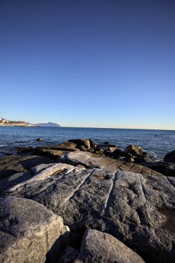 Küçük bir körfezden görünen ve dalgakıran kayaları tarafından çerçevelenen binalar ve uçurumun kenarındaki bir çıkıntının ucu.