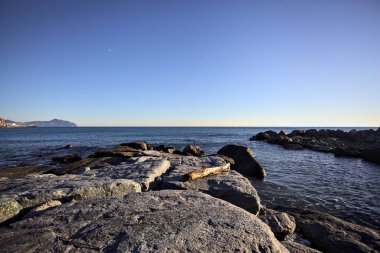 Küçük bir körfezden görünen ve dalgakıran kayaları tarafından çerçevelenen binalar ve uçurumun kenarındaki bir çıkıntının ucu.