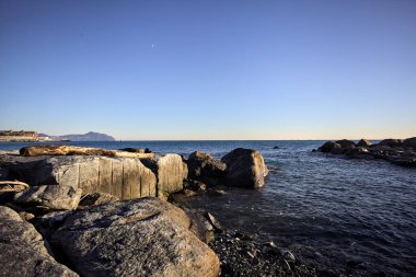 Küçük bir körfezden görünen ve dalgakıran kayaları tarafından çerçevelenen binalar ve uçurumun kenarındaki bir çıkıntının ucu.