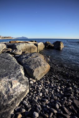 Tip of a promontory with buildings and a cliff away in the distance seen from a small cove and framed by rocks of a breakwater