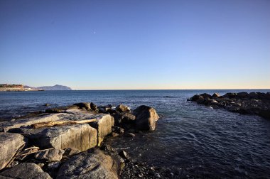 Küçük bir körfezden görünen ve dalgakıran kayaları tarafından çerçevelenen binalar ve uçurumun kenarındaki bir çıkıntının ucu.