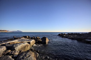 Küçük bir körfezden görünen ve dalgakıran kayaları tarafından çerçevelenen binalar ve uçurumun kenarındaki bir çıkıntının ucu.