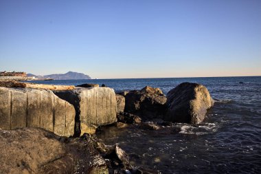 Küçük bir körfezden görünen ve dalgakıran kayaları tarafından çerçevelenen binalar ve uçurumun kenarındaki bir çıkıntının ucu.