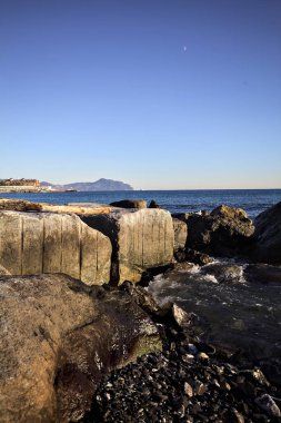 Tip of a promontory with buildings and a cliff away in the distance seen from a small cove and framed by rocks of a breakwater