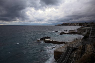 Dalgalar, alacakaranlıkta bulutlu bir günde, küçük bir koydan engebeli bir denize doğru uzanır. Üzerlerindeki kayalıklardan görülür. Arka planda fildişi uçları vardır.