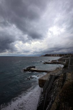 Dalgalar, alacakaranlıkta bulutlu bir günde, küçük bir koydan engebeli bir denize doğru uzanır. Üzerlerindeki kayalıklardan görülür. Arka planda fildişi uçları vardır.