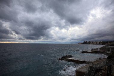 Dalgalar, alacakaranlıkta bulutlu bir günde, küçük bir koydan engebeli bir denize doğru uzanır. Üzerlerindeki kayalıklardan görülür. Arka planda fildişi uçları vardır.