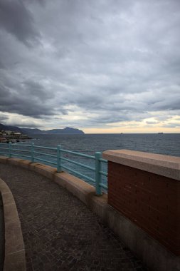 Alacakaranlıkta bulutlu bir günde deniz, uzakta uçurumlar, yolda mavi raylı bir kaldırımdan görülür.
