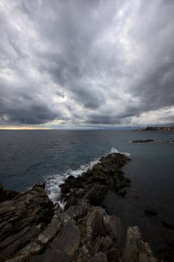 Alacakaranlıkta bulutlu bir günde, bir uçurumun kayaları ve dalgaların altında ezildiği dalgalar.