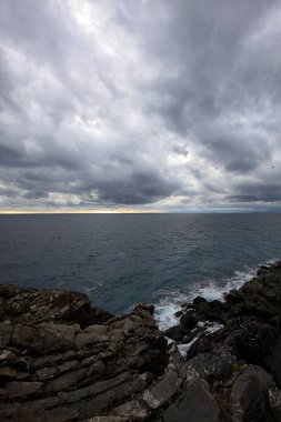 Alacakaranlıkta bulutlu bir günde, bir uçurumun kayaları ve dalgaların altında ezildiği dalgalar.