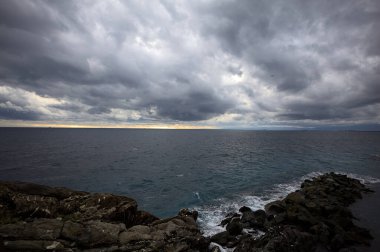 Alacakaranlıkta bulutlu bir günde, bir uçurumun kayaları ve dalgaların altında ezildiği dalgalar.