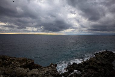 Alacakaranlıkta bulutlu bir günde, bir uçurumun kayaları ve dalgaların altında ezildiği dalgalar.