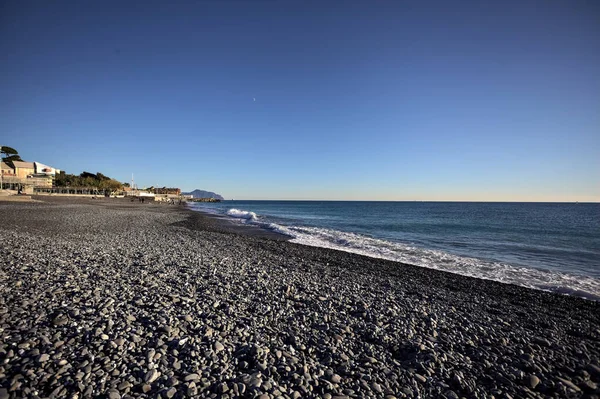 Foreshore with crushing waves on it on a sunny day, with buildings and a cliff in the distance, counterlit by the sun