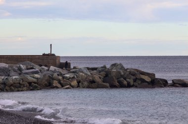 Gün batımında, bulutlu bir günde kayalıklarda duran bir balıkçıyla bir kumsalın yanında dalgalanan su.