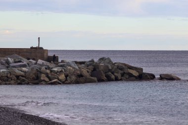 Gün batımında, bulutlu bir günde kayalıklarda duran bir balıkçıyla bir kumsalın yanında dalgalanan su.