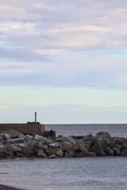 Gün batımında, bulutlu bir günde kayalıklarda duran bir balıkçıyla bir kumsalın yanında dalgalanan su.