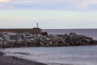 Gün batımında, bulutlu bir günde kayalıklarda duran bir balıkçıyla bir kumsalın yanında dalgalanan su.