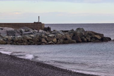 Gün batımında, bulutlu bir günde kayalıklarda duran bir balıkçıyla bir kumsalın yanında dalgalanan su.