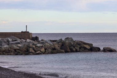 Gün batımında, bulutlu bir günde kayalıklarda duran bir balıkçıyla bir kumsalın yanında dalgalanan su.