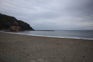 Kışın bulutlu bir günde, kumsalda bir bina, arka planda ağaçlarla kaplı bir uçurum ve uçurumun kenarında bir dalgakıran.