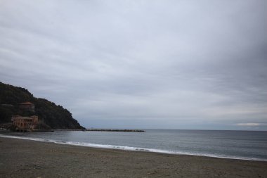 Kışın bulutlu bir günde, kumsalda bir bina, arka planda ağaçlarla kaplı bir uçurum ve uçurumun kenarında bir dalgakıran.