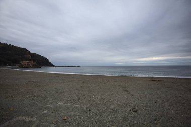 Kışın bulutlu bir günde, kumsalda bir bina, arka planda ağaçlarla kaplı bir uçurum ve uçurumun kenarında bir dalgakıran.