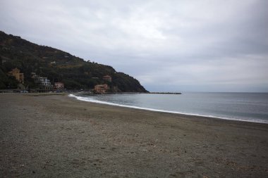 Dalgalar denize doğru uzanıyor, kapalı tatil beldeleri olan bir sahilde ve arka planda bir kasabanın binaları, kışın bulutlu bir günde.