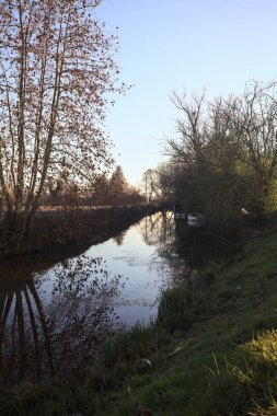 Bir tarlanın yanındaki su deresi, kıyıdaki bazı ağaçların yanında demirli tekneler, gün batımında İtalya kırsalında.