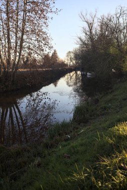 Bir tarlanın yanındaki su deresi, kıyıdaki bazı ağaçların yanında demirli tekneler, gün batımında İtalya kırsalında.