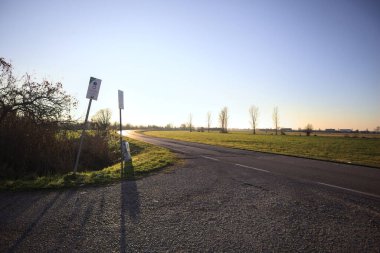 Bir tarla ve suyla çevrelenmiş bir hendekle çevrili bir yolda, asfalta dökülen güneş ışığıyla, gün batımında İtalya kırsalında