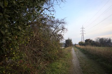 Elektrik direği ve sınırında bir elektrik hattı olan patika, gün batımında İtalya 'nın kırsal kesimindeki bir parkta, bir köyün evleriyle sınırda, ve eğlence kanalının diğer tarafında bir viyadük kanalı ile çevrili.