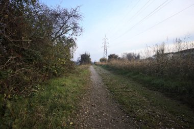 Elektrik direği ve sınırında bir elektrik hattı olan patika, gün batımında İtalya 'nın kırsal kesimindeki bir parkta, bir köyün evleriyle sınırda, ve eğlence kanalının diğer tarafında bir viyadük kanalı ile çevrili.