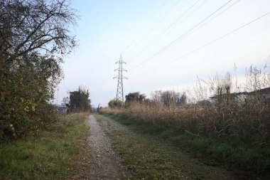 Elektrik direği ve sınırında bir elektrik hattı olan patika, gün batımında İtalya 'nın kırsal kesimindeki bir parkta, bir köyün evleriyle sınırda, ve eğlence kanalının diğer tarafında bir viyadük kanalı ile çevrili.