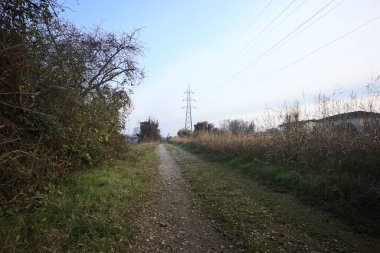Elektrik direği ve sınırında bir elektrik hattı olan patika, gün batımında İtalya 'nın kırsal kesimindeki bir parkta, bir köyün evleriyle sınırda, ve eğlence kanalının diğer tarafında bir viyadük kanalı ile çevrili.