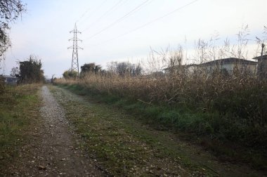 Elektrik direği ve sınırında bir elektrik hattı olan patika, gün batımında İtalya 'nın kırsal kesimindeki bir parkta, bir köyün evleriyle sınırda, ve eğlence kanalının diğer tarafında bir viyadük kanalı ile çevrili.