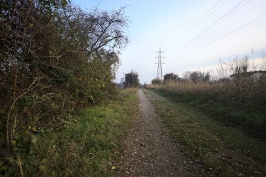 Elektrik direği ve sınırında bir elektrik hattı olan patika, gün batımında İtalya 'nın kırsal kesimindeki bir parkta, bir köyün evleriyle sınırda, ve eğlence kanalının diğer tarafında bir viyadük kanalı ile çevrili.