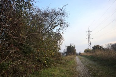 Elektrik direği ve sınırında bir elektrik hattı olan patika, gün batımında İtalya 'nın kırsal kesimindeki bir parkta, bir köyün evleriyle sınırda, ve eğlence kanalının diğer tarafında bir viyadük kanalı ile çevrili.