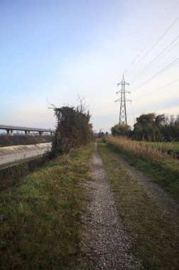 Elektrik direği ve sınırında bir elektrik hattı olan patika, gün batımında İtalya 'nın kırsal kesimindeki bir parkta, bir köyün evleriyle sınırda, ve eğlence kanalının diğer tarafında bir viyadük kanalı ile çevrili.