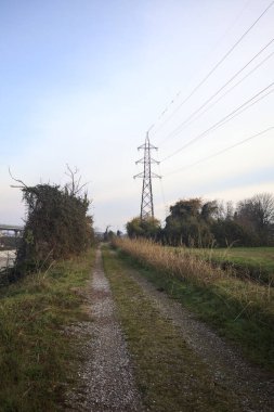 Elektrik direği ve sınırında bir elektrik hattı olan patika, gün batımında İtalya 'nın kırsal kesimindeki bir parkta, bir köyün evleriyle sınırda, ve eğlence kanalının diğer tarafında bir viyadük kanalı ile çevrili.