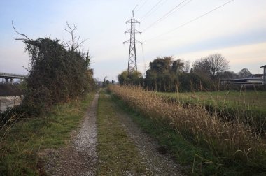 Elektrik direği ve sınırında bir elektrik hattı olan patika, gün batımında İtalya 'nın kırsal kesimindeki bir parkta, bir köyün evleriyle sınırda, ve eğlence kanalının diğer tarafında bir viyadük kanalı ile çevrili.