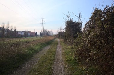 Elektrik direği ve sınırında bir elektrik hattı olan patika, gün batımında İtalya 'nın kırsal kesimindeki bir parkta, bir köyün evleriyle sınırda, ve eğlence kanalının diğer tarafında bir viyadük kanalı ile çevrili.