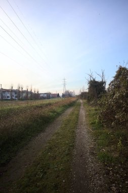 Elektrik direği ve sınırında bir elektrik hattı olan patika, gün batımında İtalya 'nın kırsal kesimindeki bir parkta, bir köyün evleriyle sınırda, ve eğlence kanalının diğer tarafında bir viyadük kanalı ile çevrili.