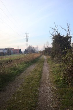 Trail with an electricity mast and an overhead powerline at its edge, in a park in the italian countryside at sunset, bordered by houses of a village, and with a viaduct on the other side of a diversionary channel