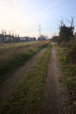 Trail with an electricity mast and an overhead powerline at its edge, in a park in the italian countryside at sunset, bordered by houses of a village, and with a viaduct on the other side of a diversionary channel