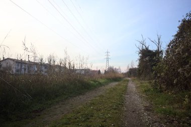 Elektrik direği ve sınırında bir elektrik hattı olan patika, gün batımında İtalya 'nın kırsal kesimindeki bir parkta, bir köyün evleriyle sınırda, ve eğlence kanalının diğer tarafında bir viyadük kanalı ile çevrili.